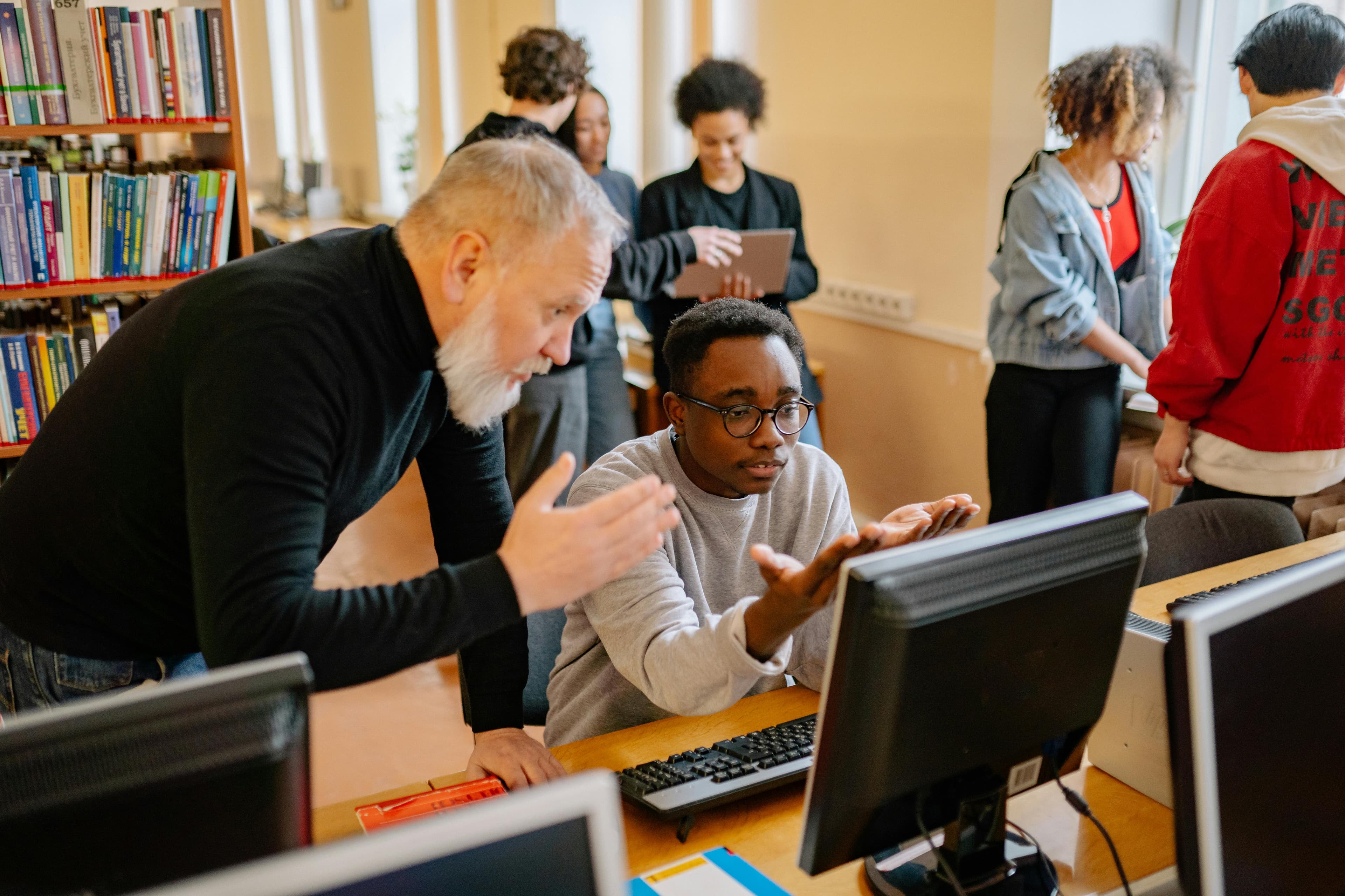 Teacher helping a student at a computer in a busy library setting, engaging in educational interaction