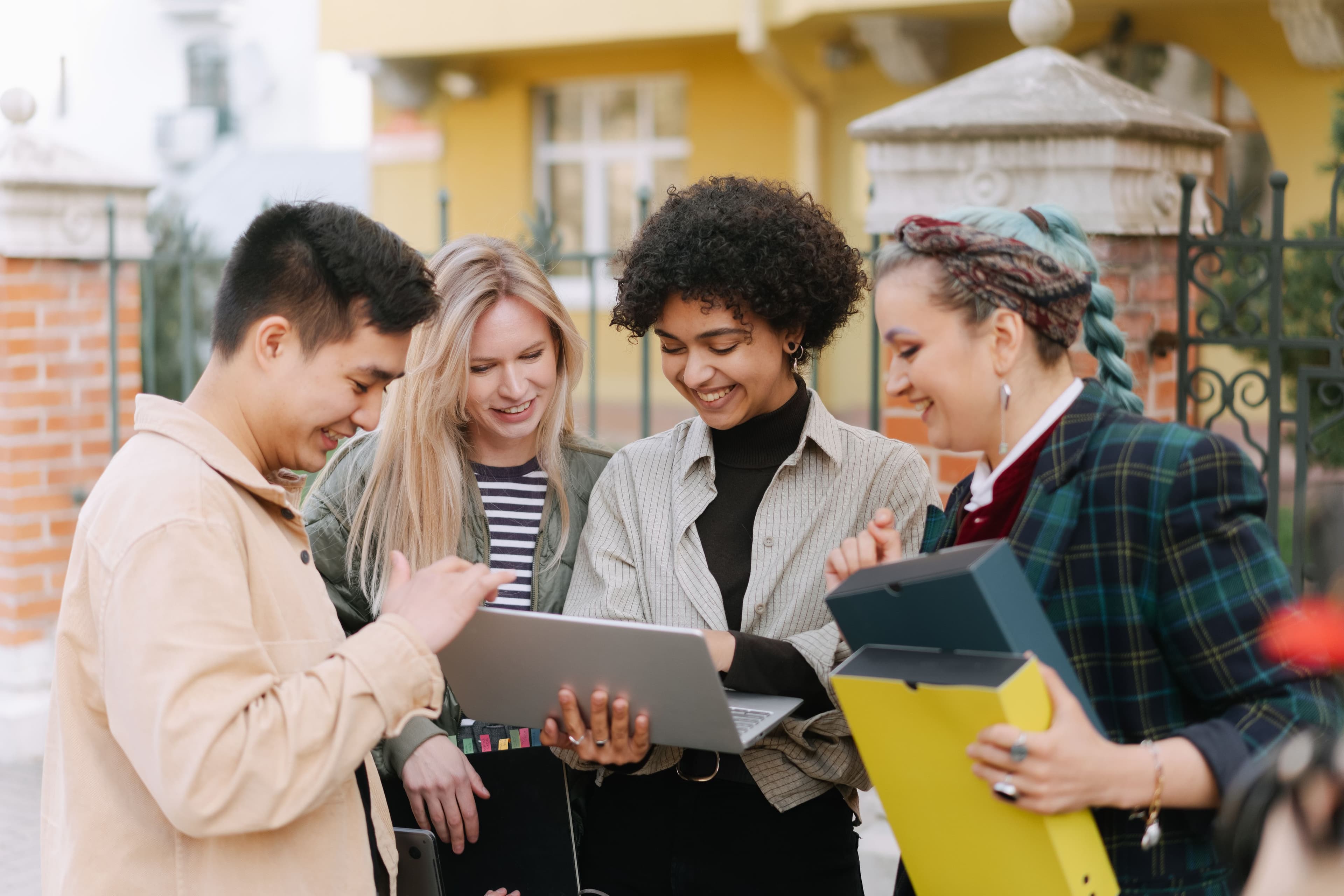 A diverse group of young adults smiling and collaborating outdoors with a laptop